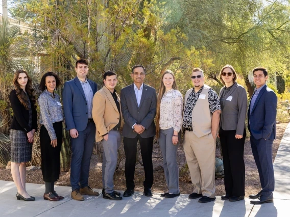 This image shows a group of nine individuals standing outdoors, posing for a photo in front of lush desert vegetation including cacti and other greenery. They are dressed in professional and business-casual attire, smiling and looking at the camera in bright daylight.